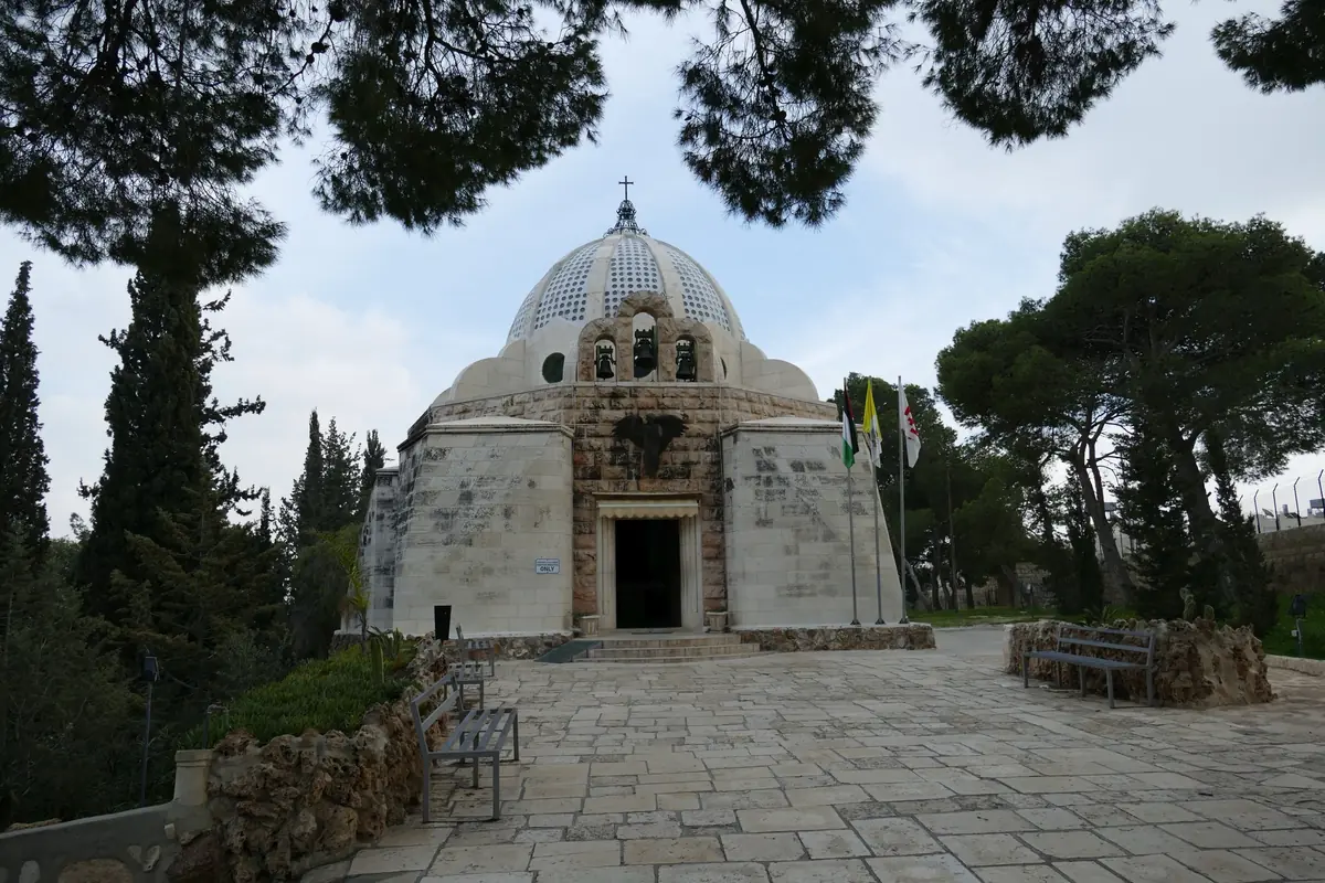 קפלה מאבן בהירה עם כיפה מחוררת, בקצה רחבה מרוצפת המוקפת עצי אורן