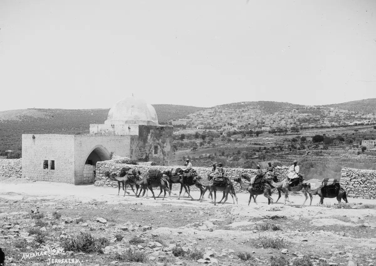 צילום ישן בשחור-לבן: מבנה לבן קטן עם כיפה לצד דרך, שיירת גמלים לפניו, גבעות וכפר ברקע