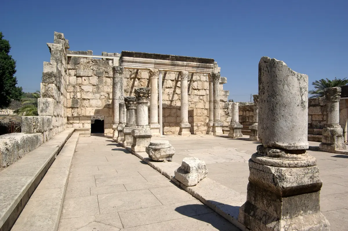חורבות בית הכנסת מאבן גיר לבנה בכפר נחום: קיר עם עמודים וכותרות, ריצוף בהיר ושברי עמודים תחת שמיים כחולים