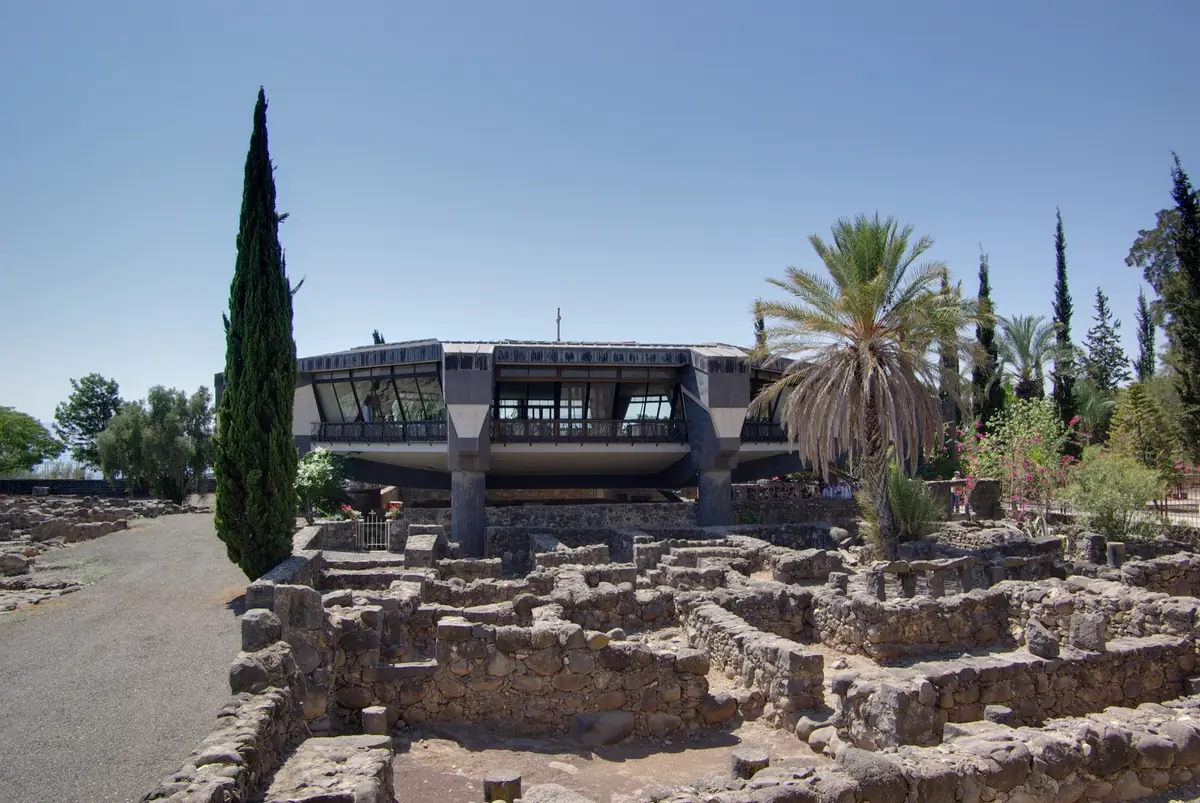 כנסייה מודרנית עם חלונות רחבים המוצבת על עמודים מעל חורבות בזלת, בין ברוש לעצי דקל