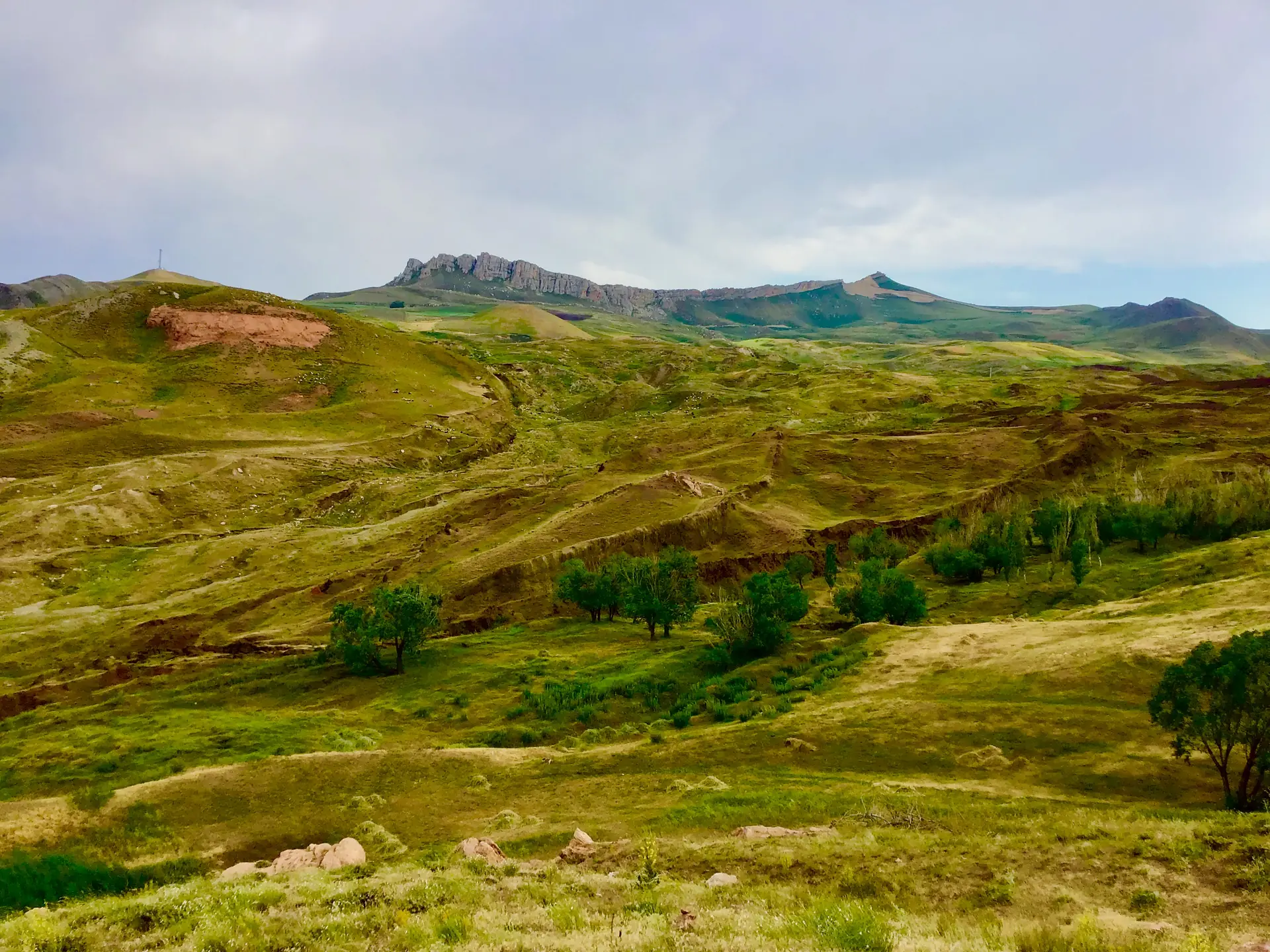 A green valley of rolling ground under a rocky ridge, with a long tree-lined mound in the middle