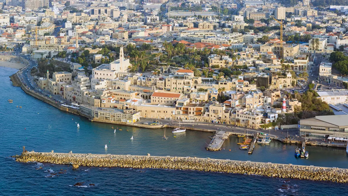 מבט אווירי על יפו העתיקה: בתים בהירים על הגבעה מתחת למגדל הפעמונים של כנסיית פטרוס, נמל קטן מאחורי שובר גלים מסלעים ומגדלון מפוספס באדום ולבן