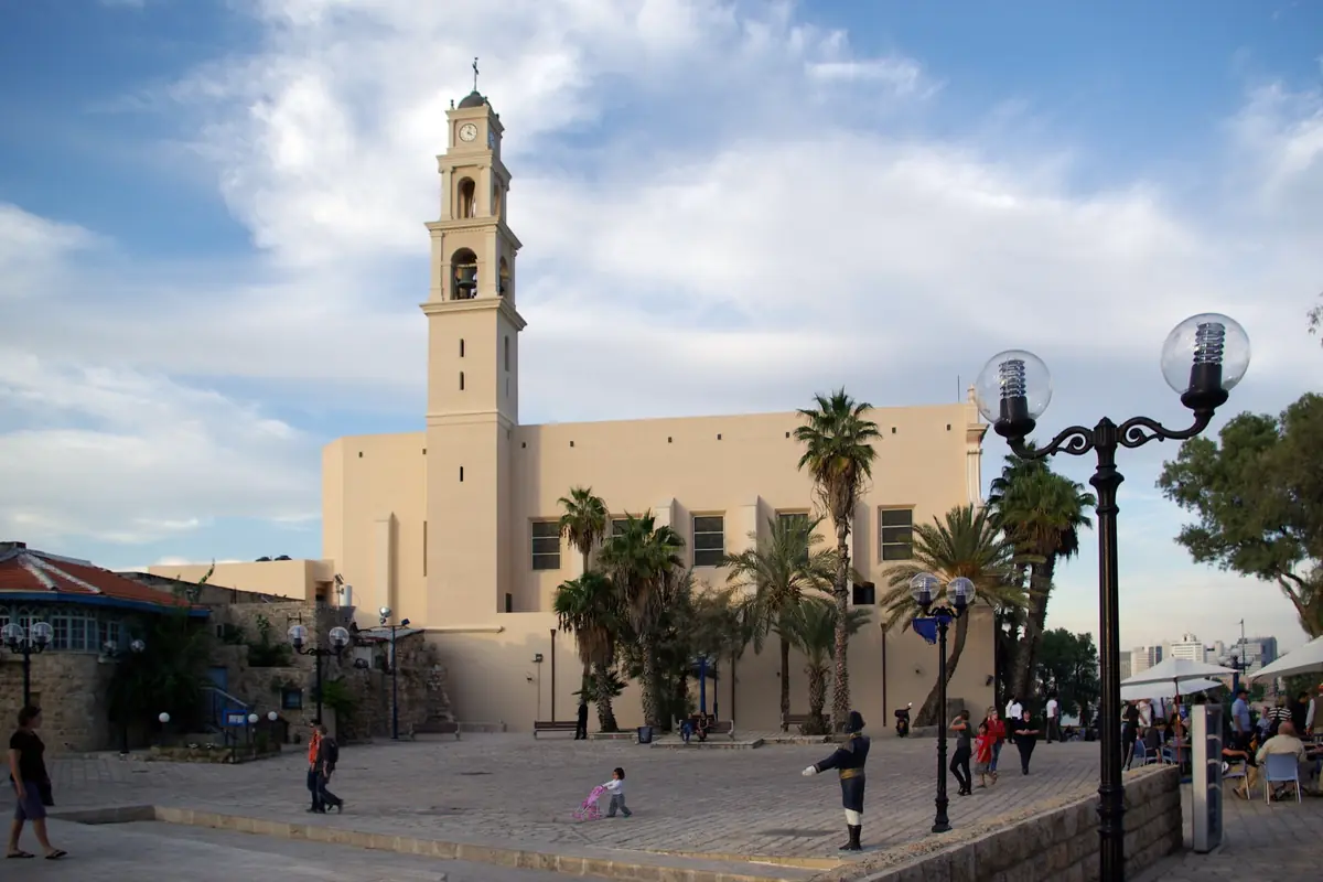כנסיית פטרוס הקדוש ביפו, חזית בהירה ומגדל פעמונים גבוה, בקצה כיכר מרוצפת עם דקלים ופנסי רחוב