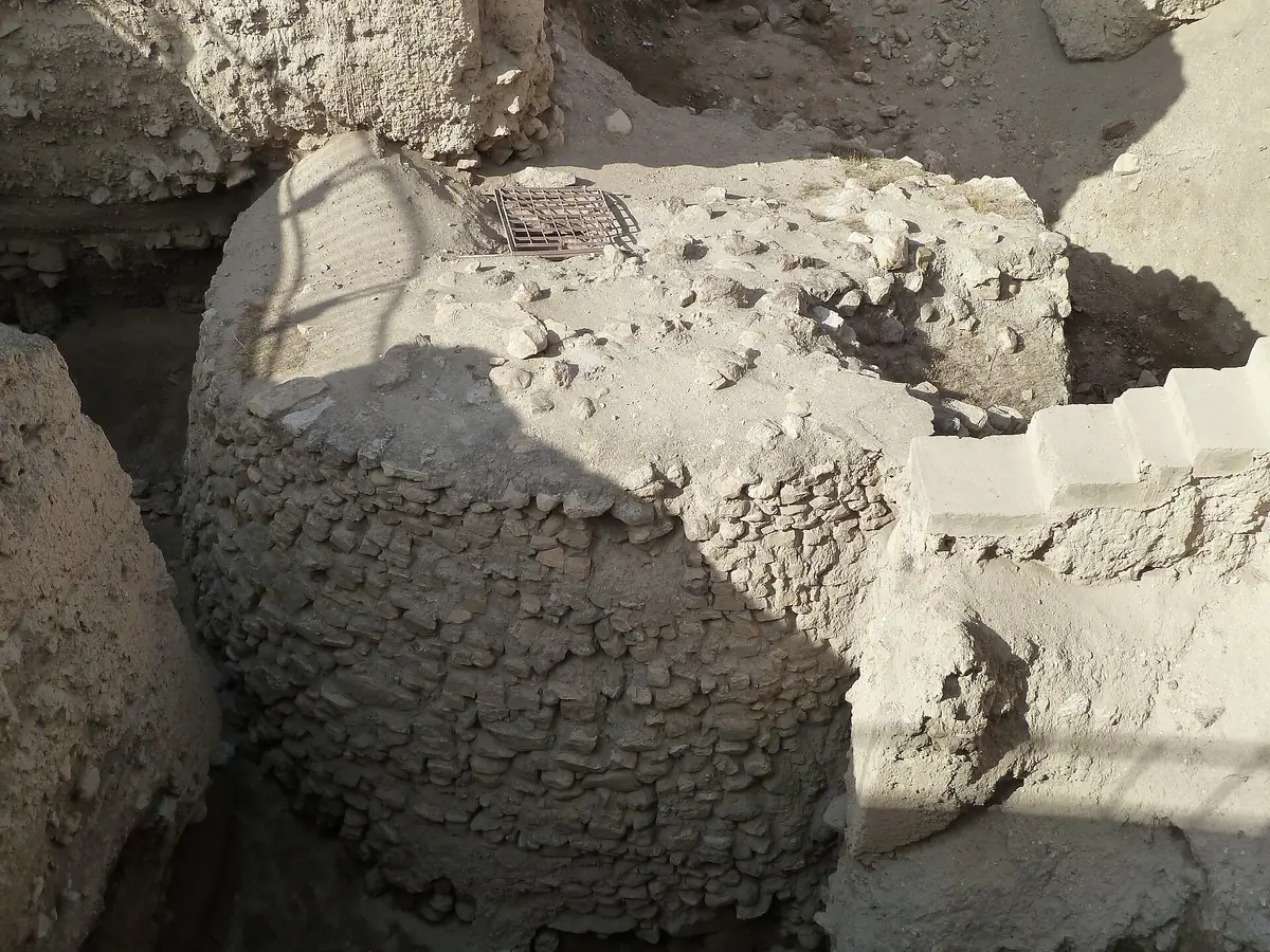 A round dry-stone tower at the bottom of an excavation trench, with a grille laid on top