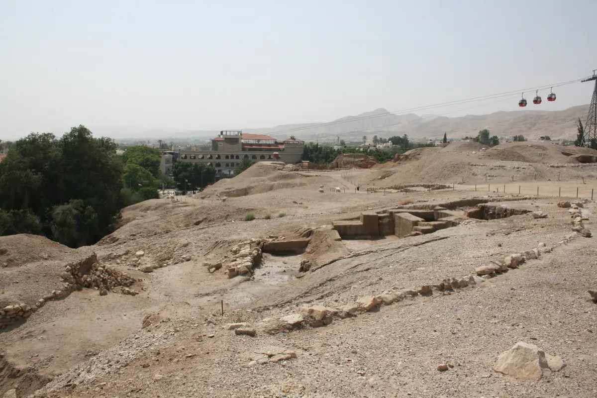 Excavated remains on the arid top of Tell es-Sultan, with red cable-car cabins and mountains in the background