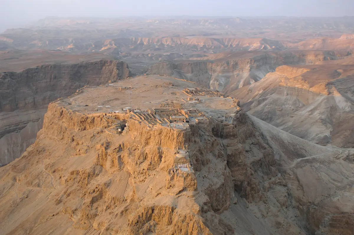 מבט אווירי על רמת מצדה: פסגה שטוחה מכוסה חורבות, מצוקים זהובים וערוצי מדבר למטה