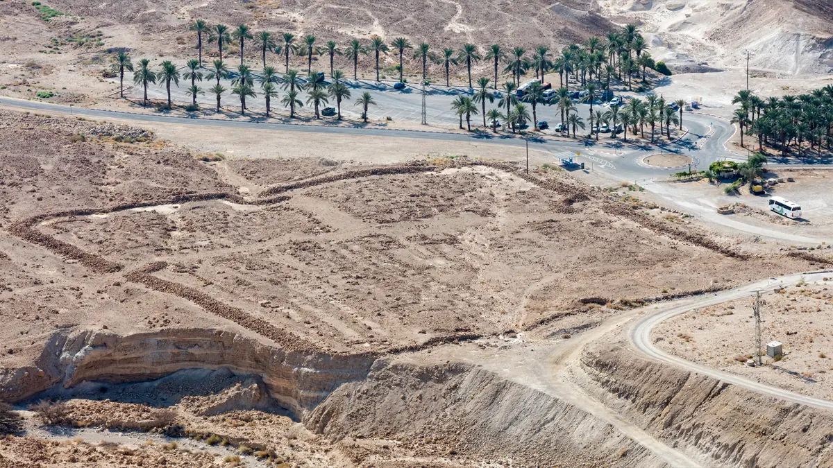 מתחם מלבני מאבני גוויל במדבר למרגלות מצדה, ומאחוריו חניון מוקף דקלים