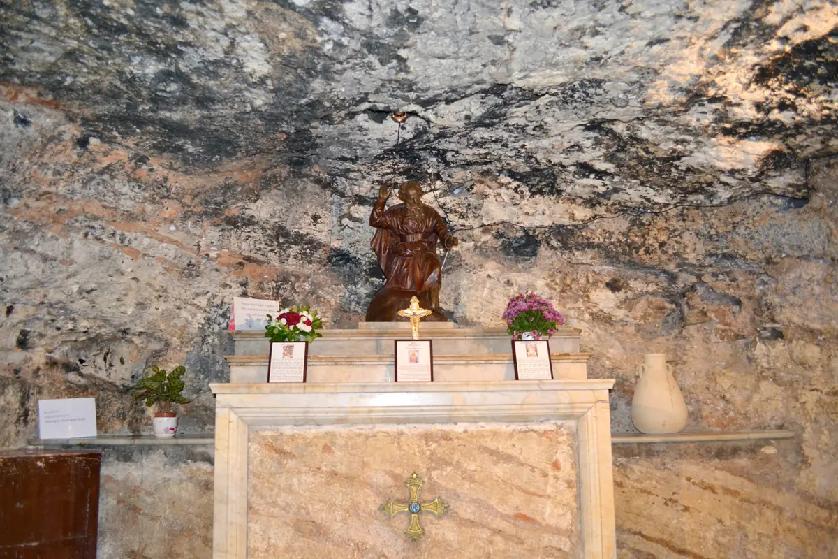 Small marble altar in a cave with a rock ceiling, topped by a wooden statuette of Elijah, with flowers and a jar