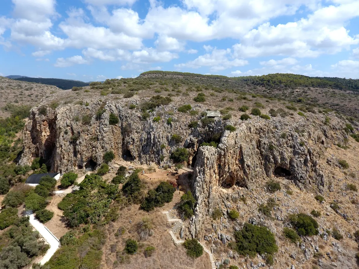 Aerial view of a limestone cliff pierced by large cave openings, with a stepped path and scrubland vegetation