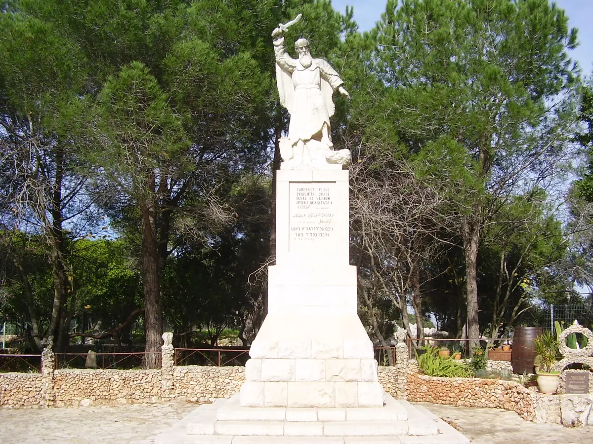 White statue of a bearded prophet brandishing a sword over a fallen man, on a tall inscribed pedestal surrounded by pine trees