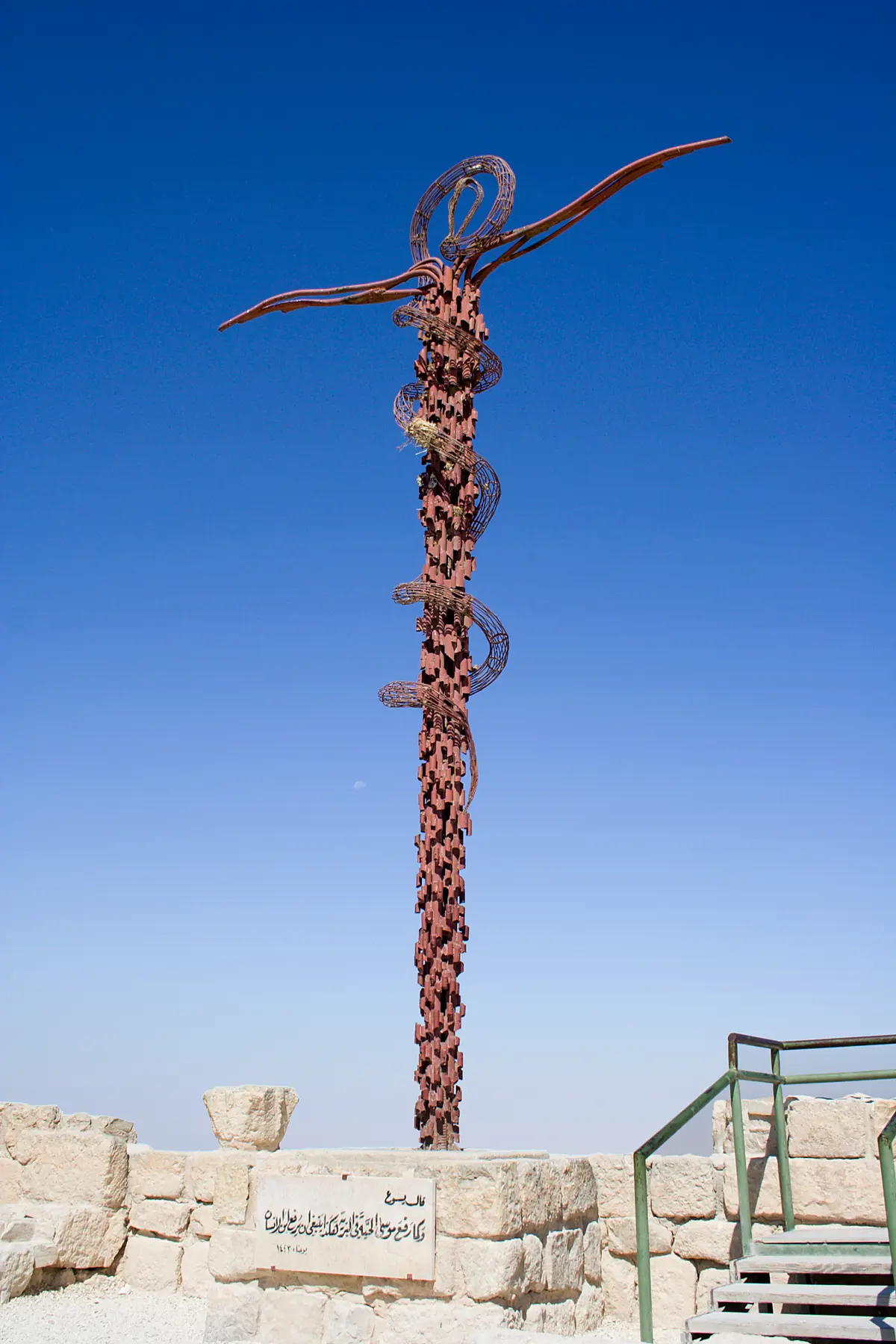 Grande sculpture en métal rouille représentant un serpent enroulé autour d'une croix, sur fond de ciel bleu