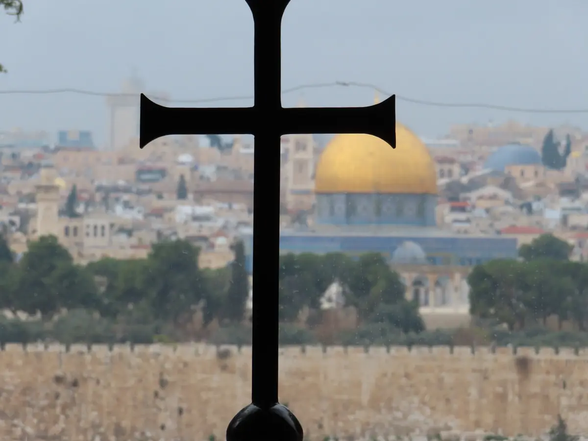 Black cross silhouetted in a window, with the golden Dome of the Rock and the Old City walls in the background