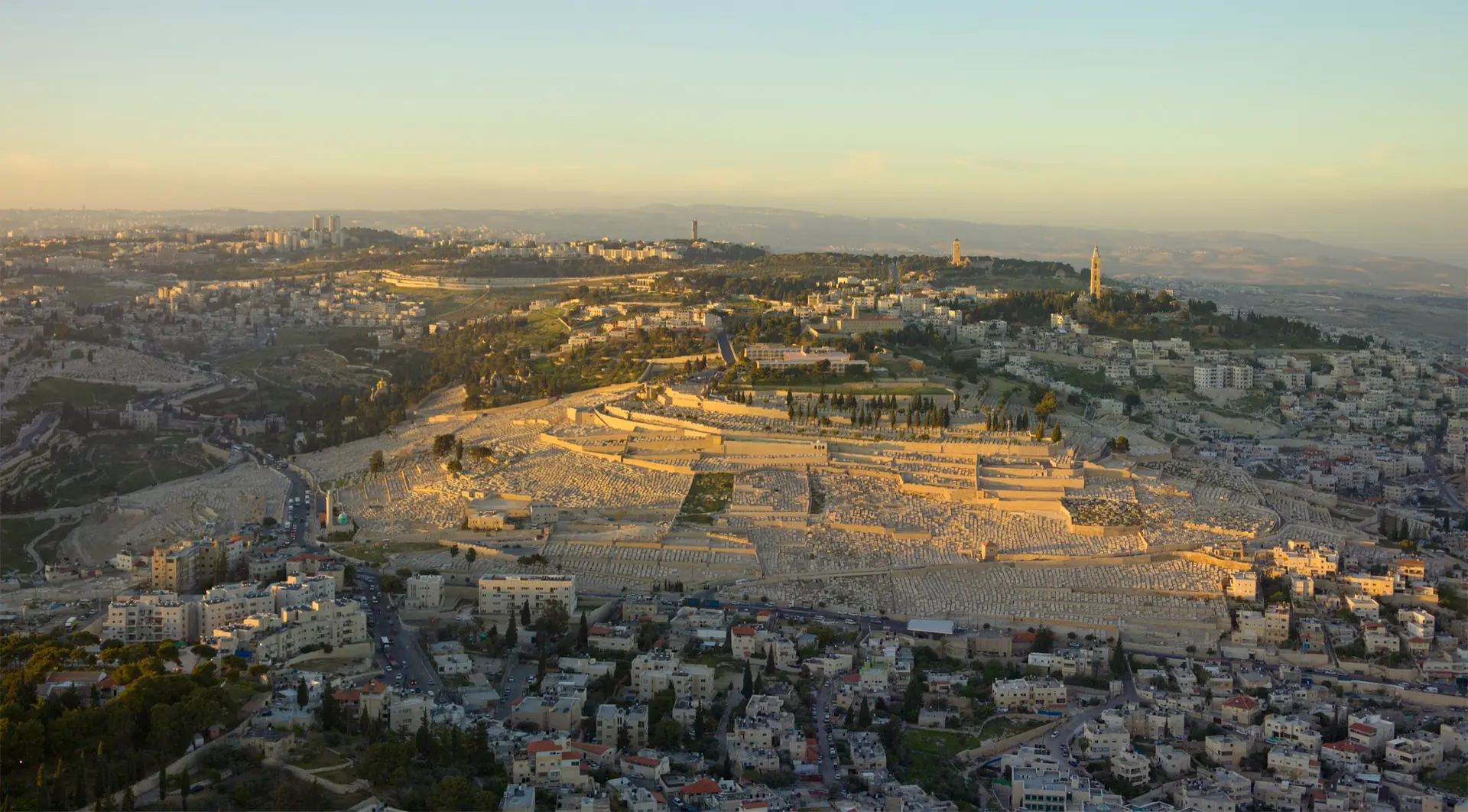 Aerial view of the Mount of Olives at sunset: the slope covered with the white tombs of the Jewish cemetery, built-up neighbourhoods around it and two bell towers on the ridge