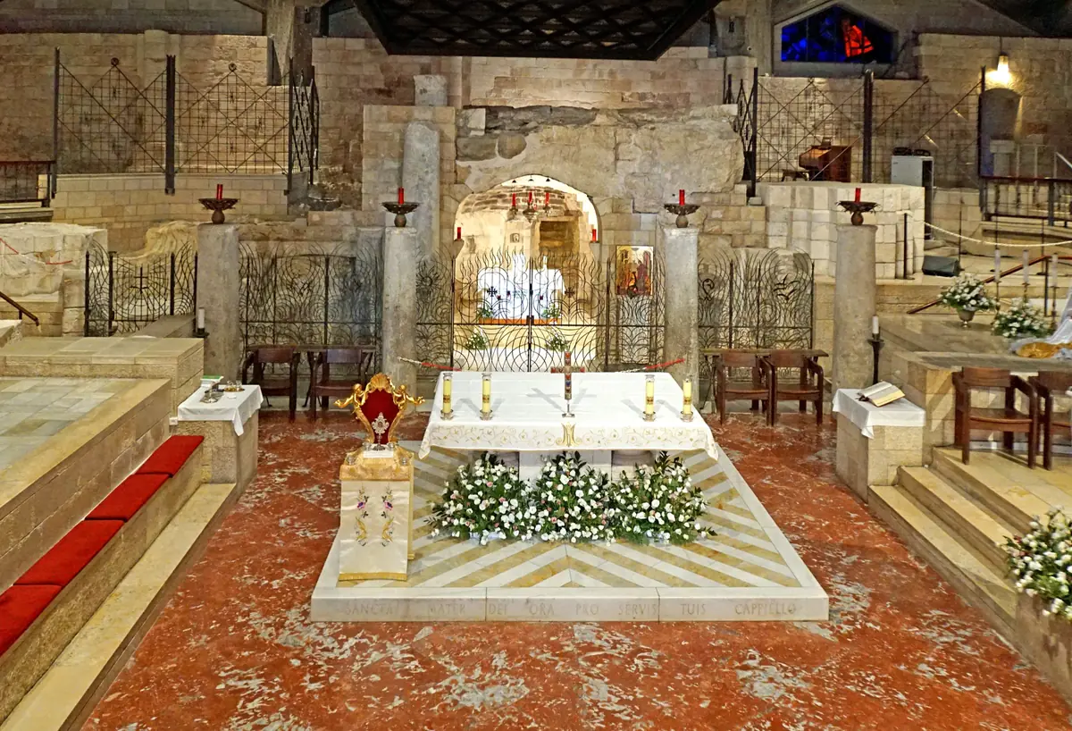מזבח שיש לבן מקושט בפרחים לפני מערה חצובה בסלע הסגורה בסורג, בין עמודים עתיקים