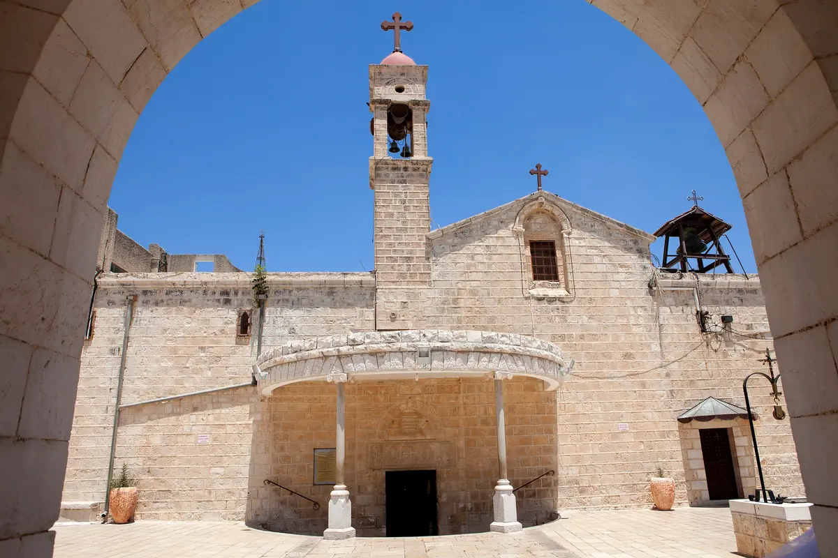 חזית כנסייה מאבן בהירה עם מגדל פעמונים ומרפסת מקושתת, במבט דרך קשת
