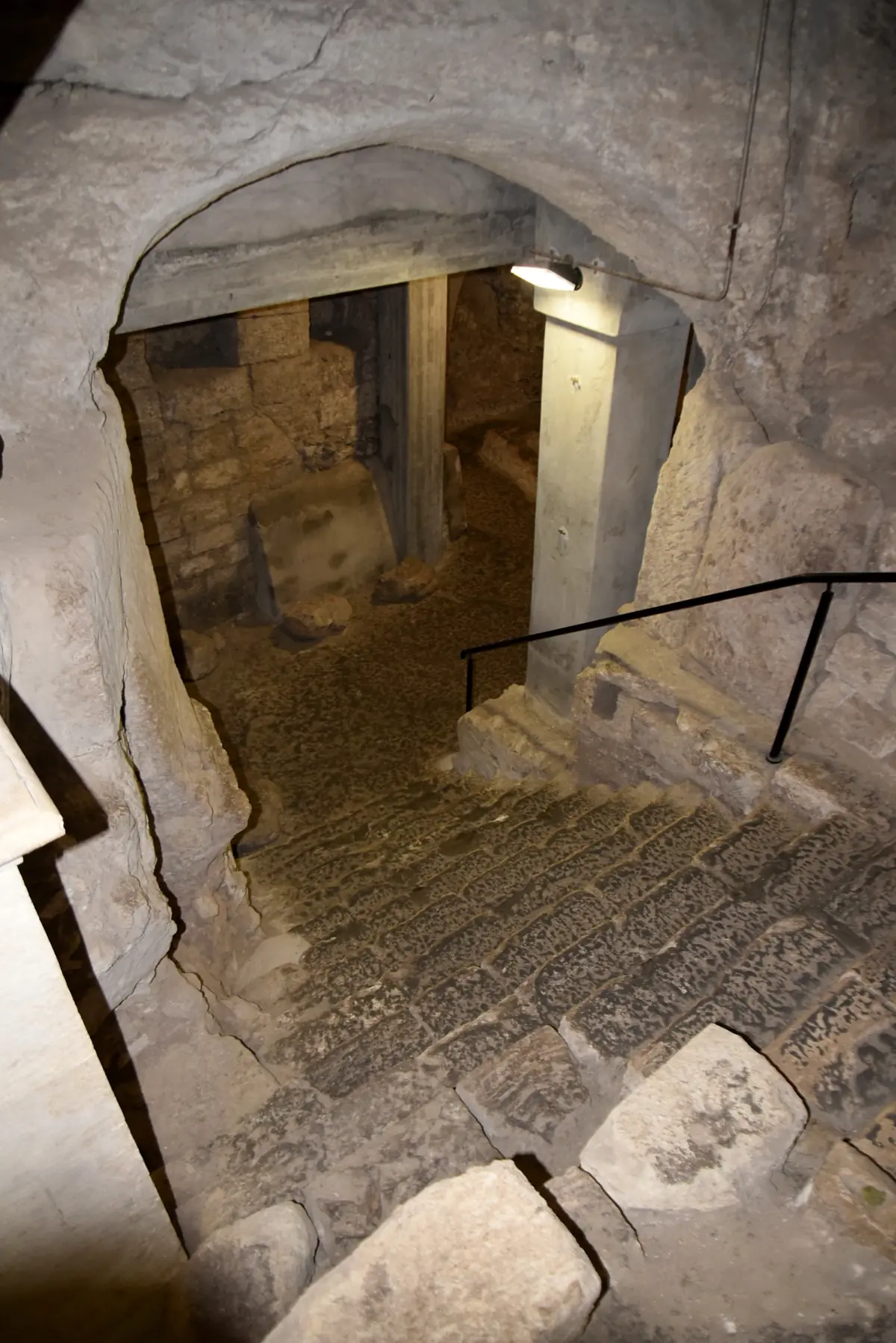 גרם מדרגות אבן שחוק היורד מתחת לקשת חצובה בסלע אל חלל תת-קרקעי מואר