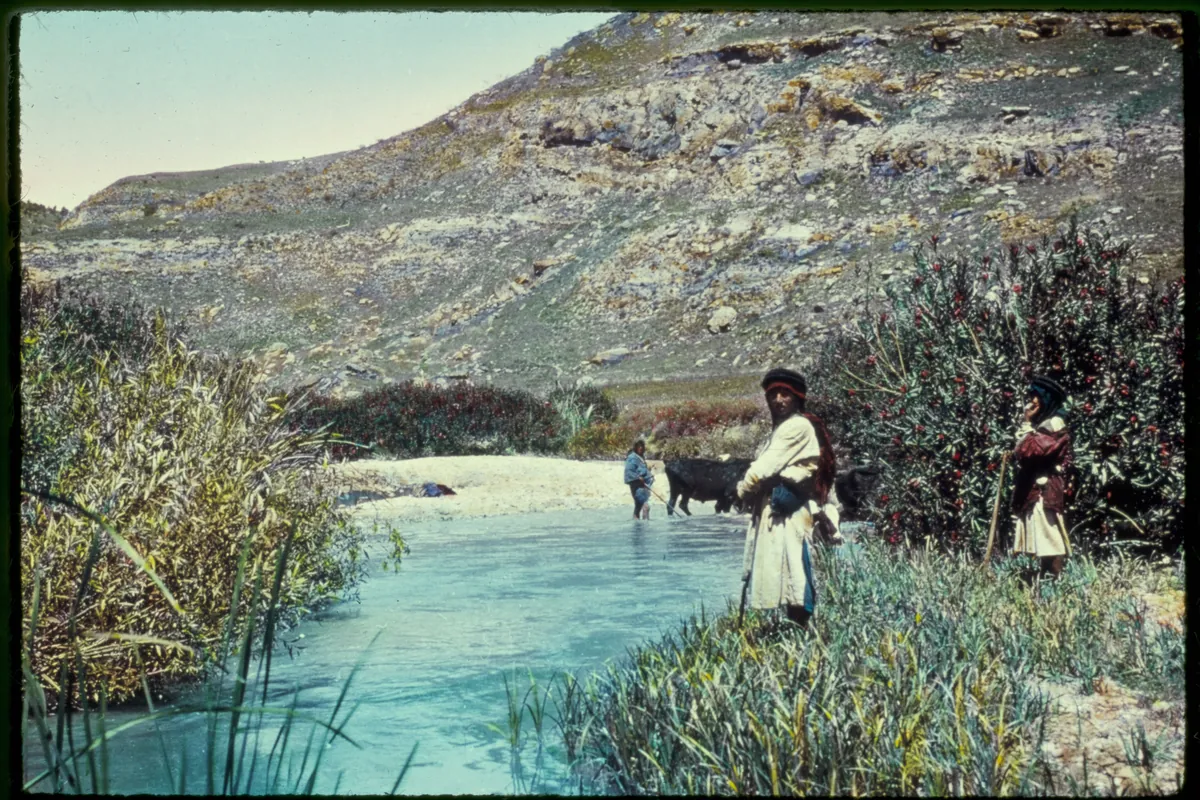 Photo ancienne colorisée : une rivière entre les roseaux au pied de collines, avec deux hommes sur la rive