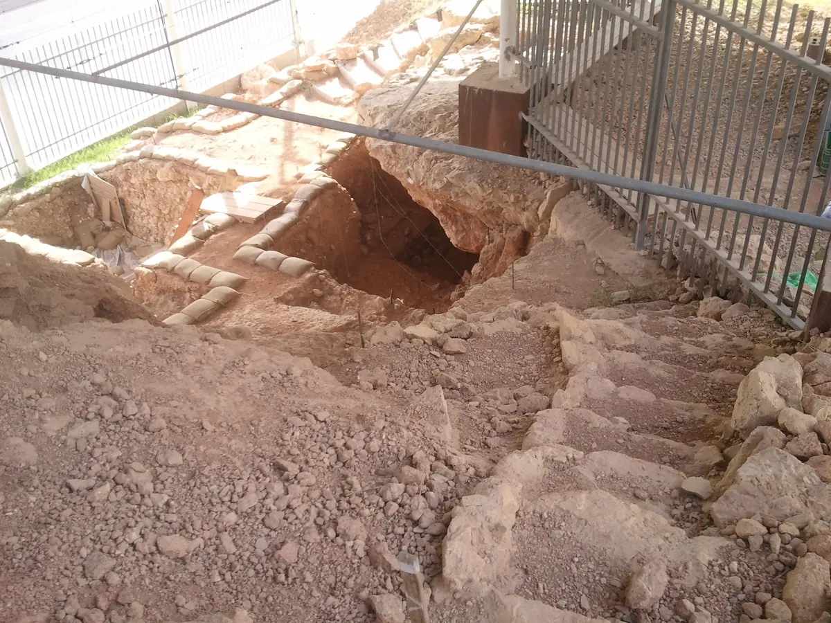 שטח חפירה מדורג בתוך משקע אדמדם, מאחורי גדר מתכת