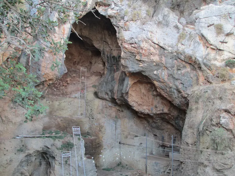 Vue aérienne d'une falaise calcaire percée de plusieurs grottes, au pied de laquelle un sentier en escalier monte à travers la garrigue