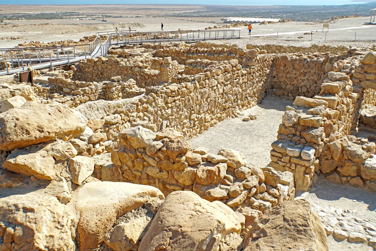 קירות אבן של חדרים מלבניים חפורים, עם שביל מבקרים מעץ ומישור ים המלח ברקע