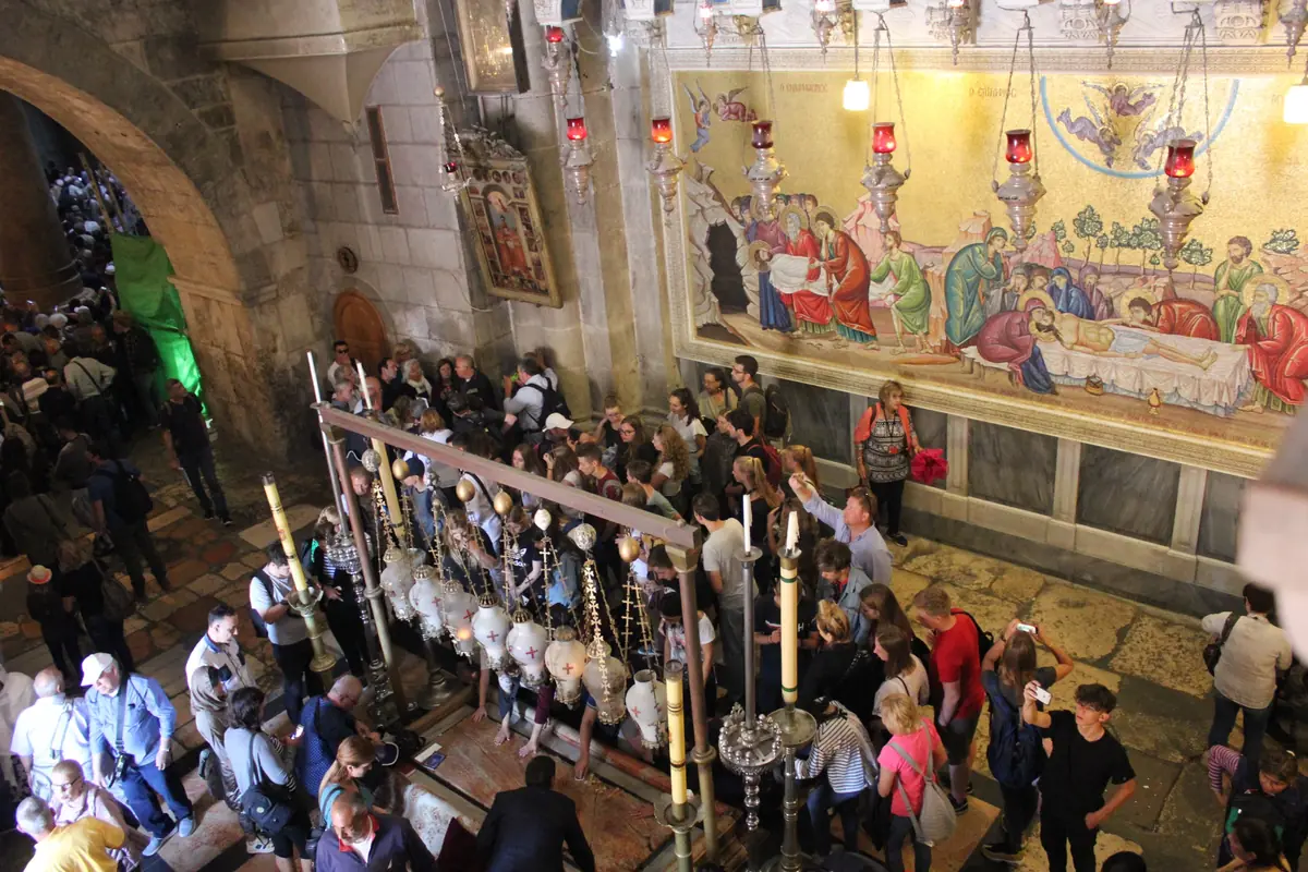 צליינים רכונים על לוח אבן מוקף מנורות תלויות, מול פסיפס מוזהב גדול המתאר את קבורת ישו