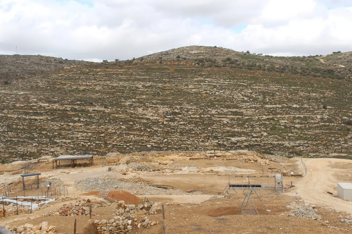 A flat excavation terrace north of Tel Shiloh, with shelters and fences, facing a terraced hill