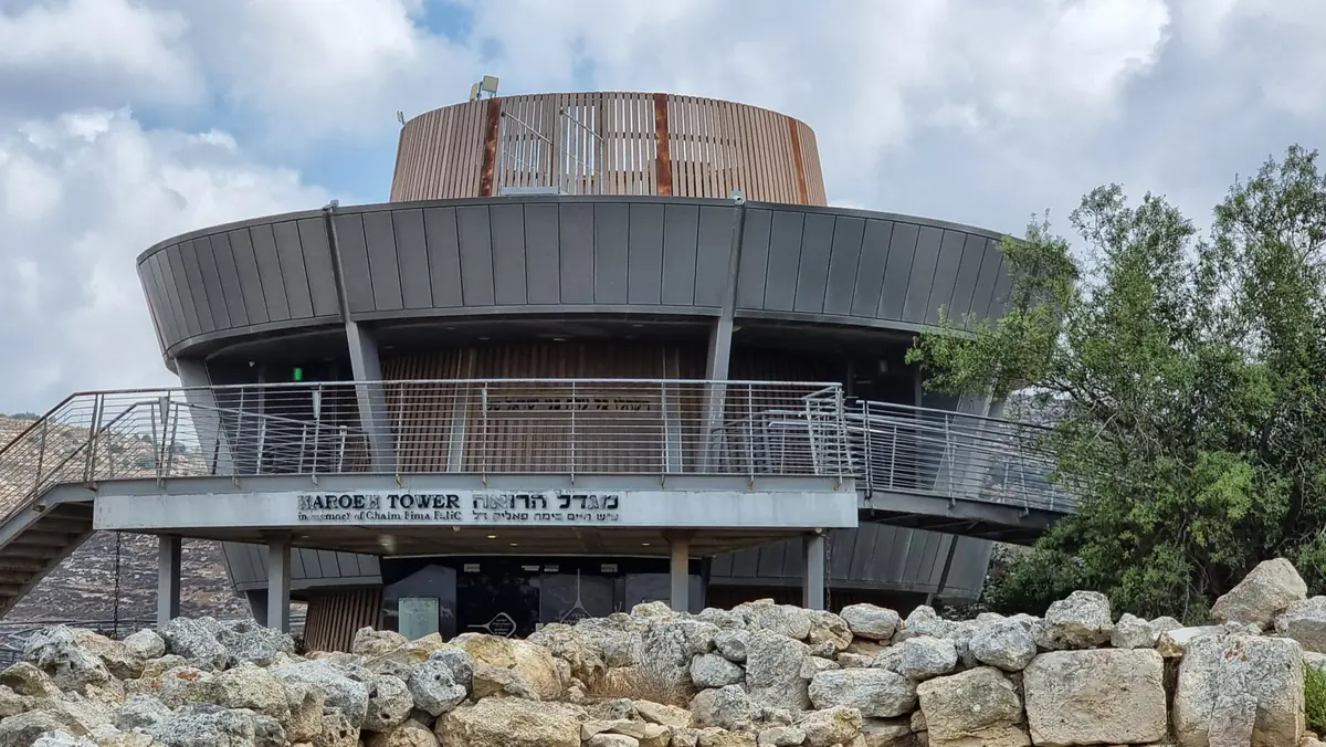 A modern circular building with a wooden top and a metal walkway, bearing the sign "HaRoeh Tower"