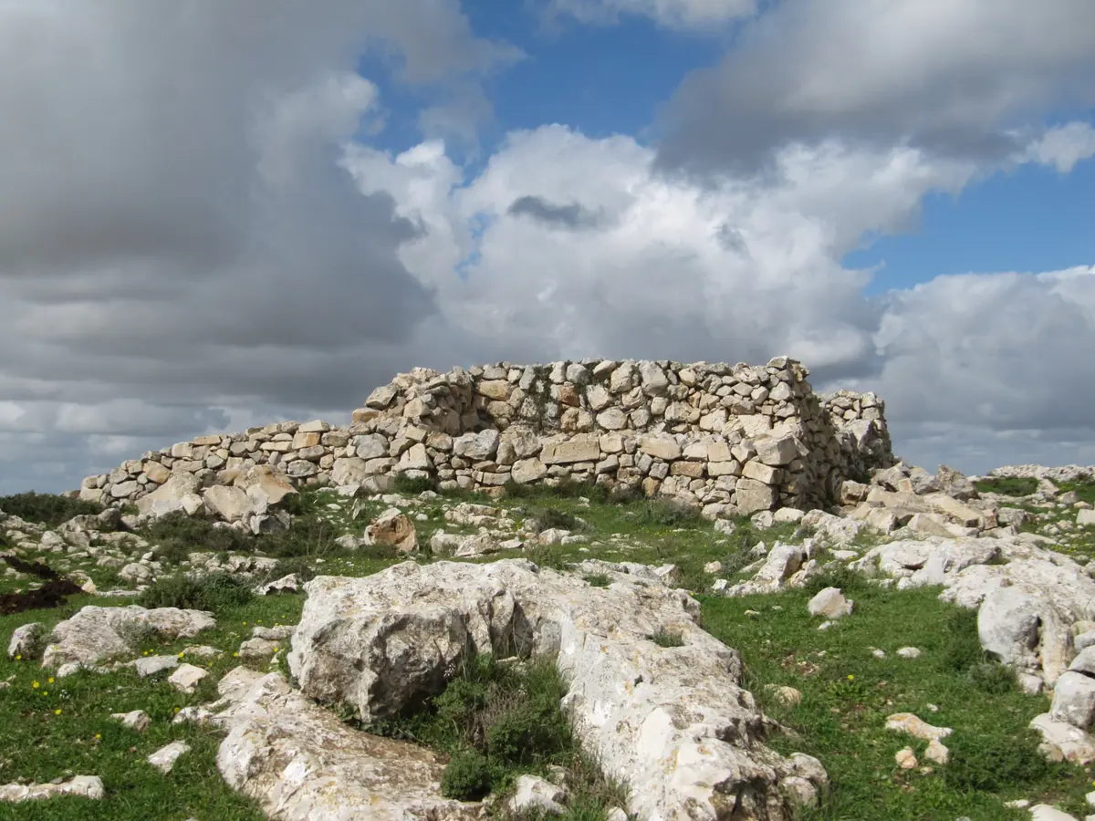A rectangular dry-stone wall on a rocky, grassy hilltop under a cloudy sky